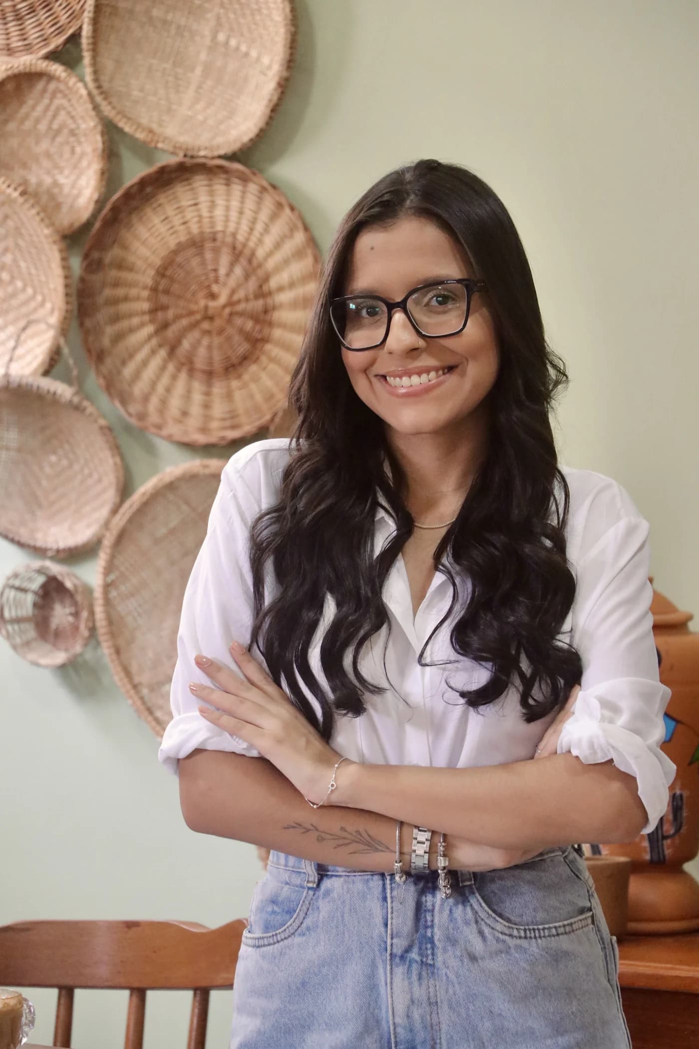 Isabella Luísa de óculos, sorrindo com os braços cruzados diante de uma parede com cestos artesanais.