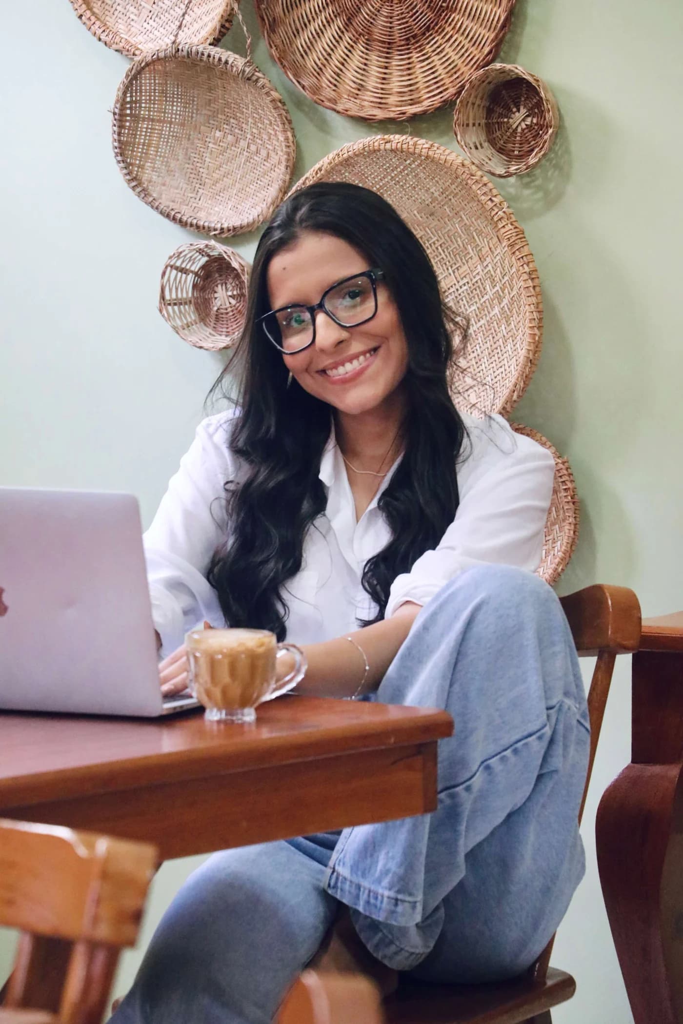 Isabella Luísa, de óculos e camisa lilás, sentada à mesa com seu notebook em um ambiente de tons naturais.